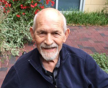 An older man with a beard sitting in a garden.
