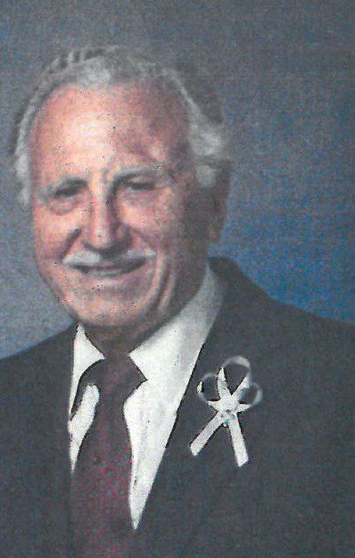 An older man in a suit and tie with a white ribbon.