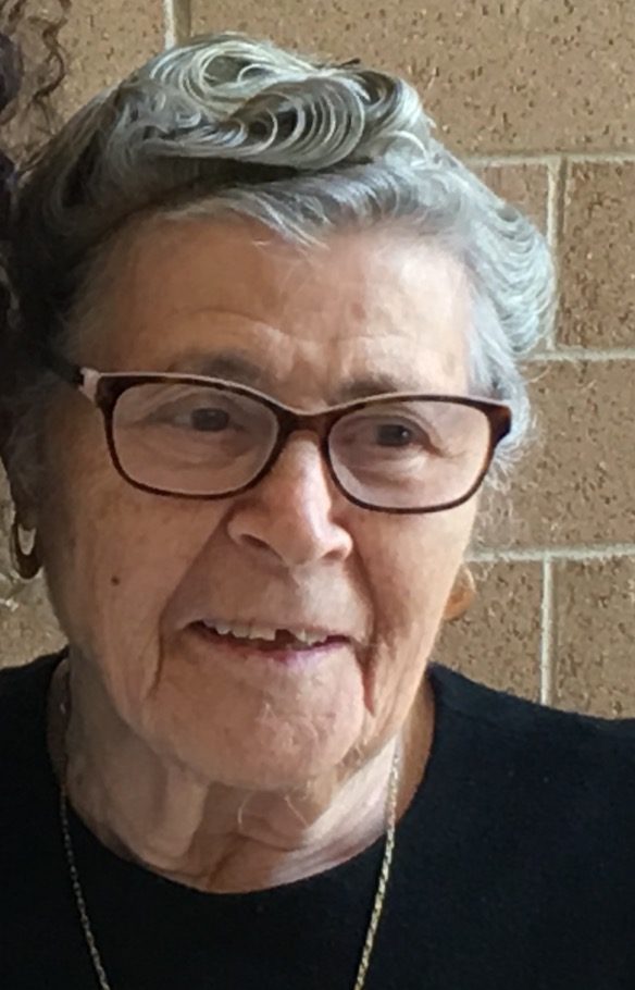 An older woman with glasses is smiling next to a brick wall.
