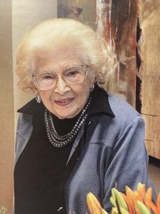 An older woman with glasses is smiling in front of a vase of flowers.