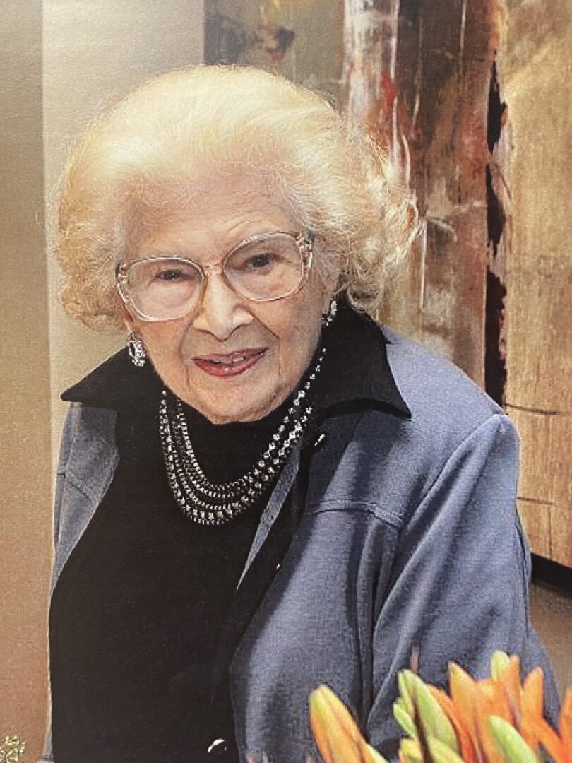 An older woman with glasses is smiling in front of a vase of flowers.