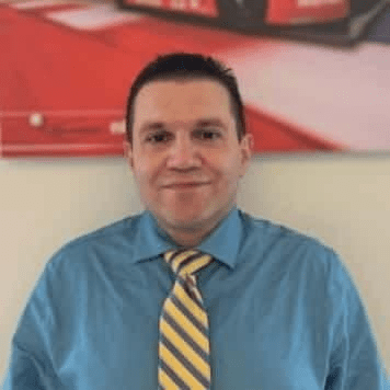 A man in a blue shirt and yellow tie standing in front of a car.