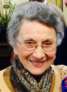 An older woman with glasses smiles in front of a group of people.