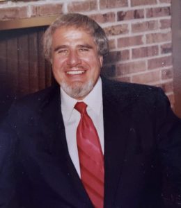 A man in a suit and tie smiling in front of a brick fireplace.