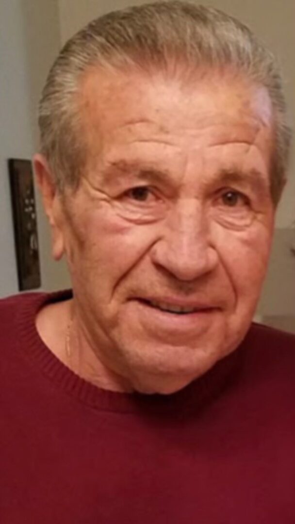 An older man in a maroon sweater is smiling.