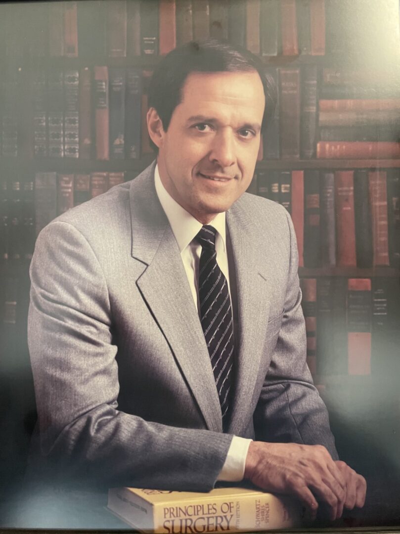 A man in a suit sits in front of a bookcase.