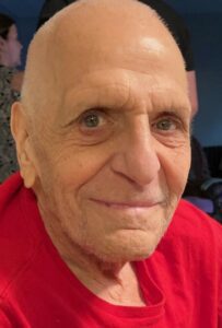 An older man in a red shirt smiles for the camera.