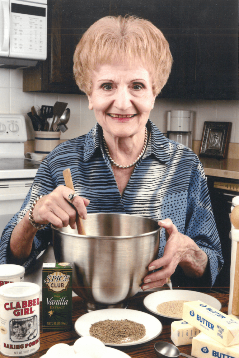 An elderly woman smiling at the camera while stirring a pot in a kitchen setting.
