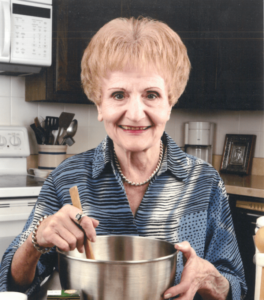An elderly woman smiling at the camera while stirring a pot in a kitchen setting.
