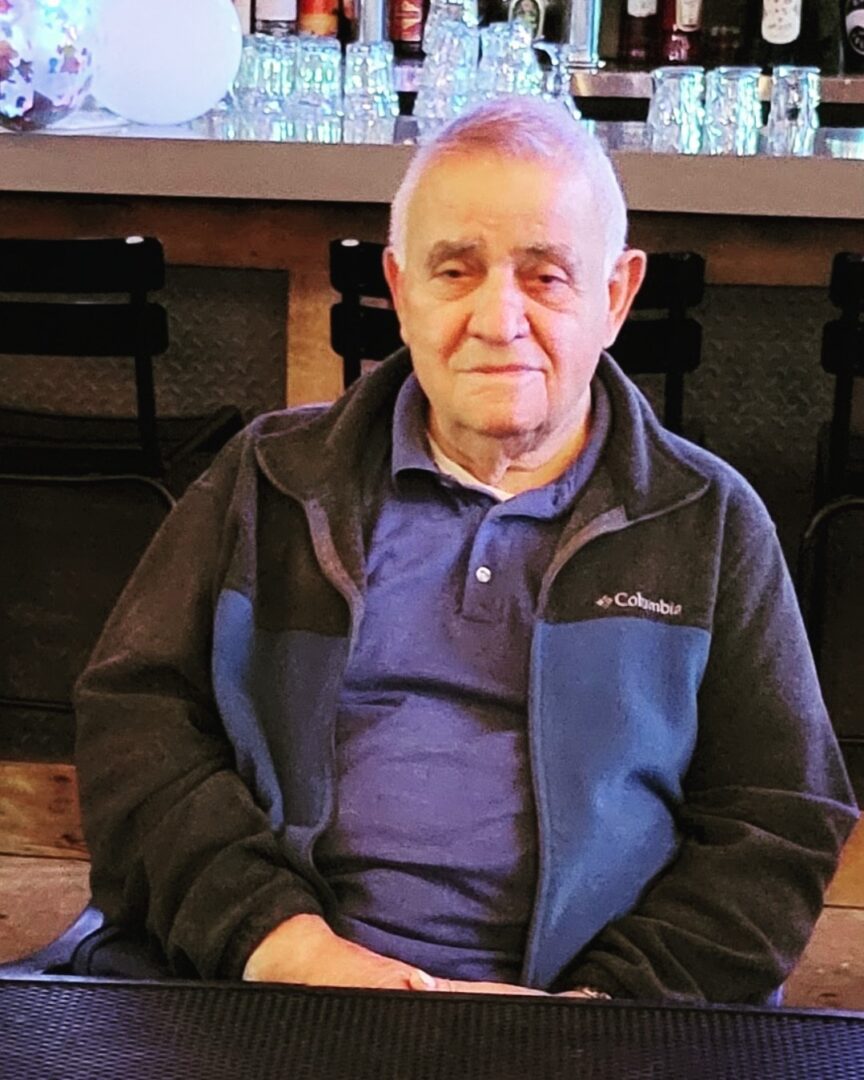 An elderly man wearing a columbia jacket sitting at a bar with shelves of bottles in the background.