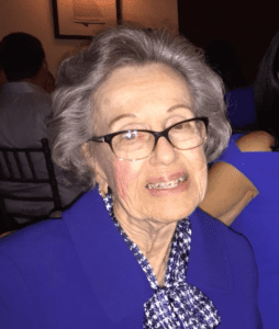 An elderly woman wearing glasses and a blue outfit smiles at the camera in a room with dim lighting.