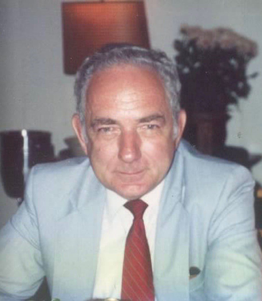 Elderly gentleman seated, wearing a light blue suit and red tie, with a serious expression.