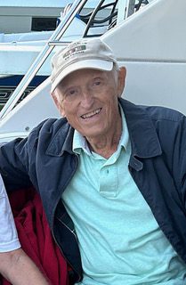 Elderly man smiling in front of a boat, wearing a cap and a jacket.