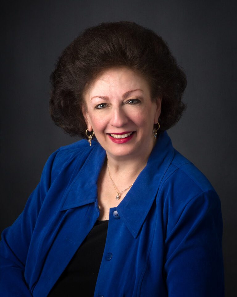 A portrait of a smiling woman with voluminous hair, wearing a blue blazer and a necklace.