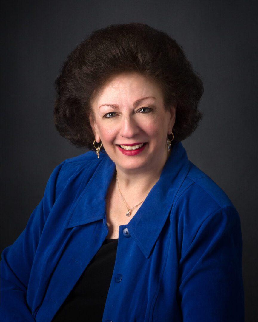 A portrait of a smiling woman with voluminous hair, wearing a blue blazer and a necklace.