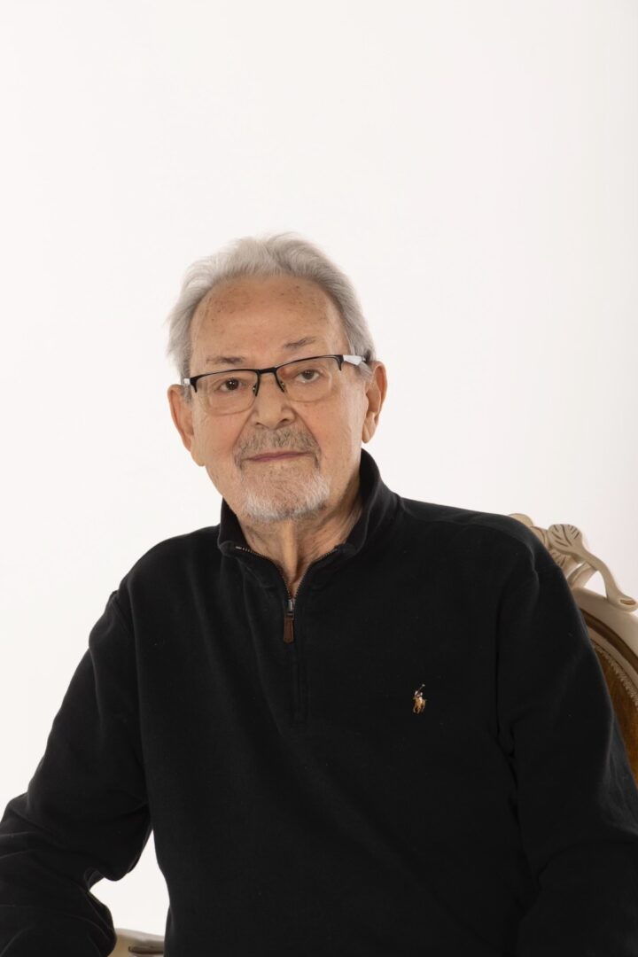 An elderly man with white hair and glasses, wearing a black zip-up sweater, sitting in front of a white background.