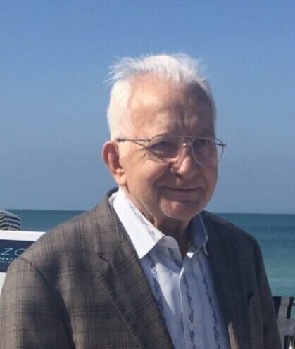 Elderly gentleman with glasses smiling at the camera, seaside in the background.