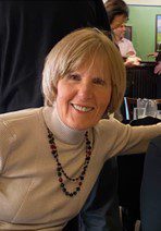 A smiling woman with short hair wearing a turtleneck and a necklace inside a room.
